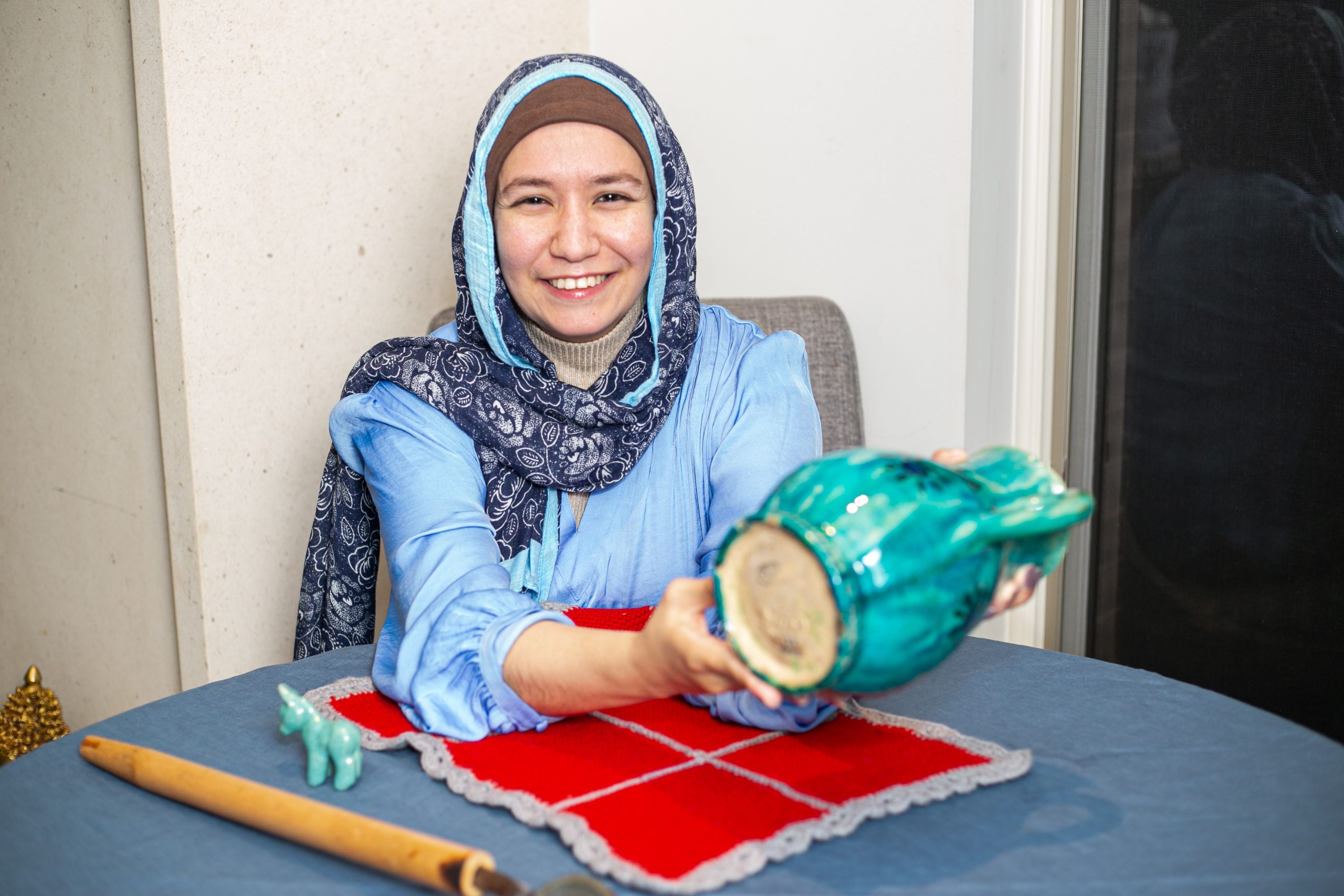 A smiling woman wearing a blue hijab and shirt holds up a turquoise ceramic pitcher.
