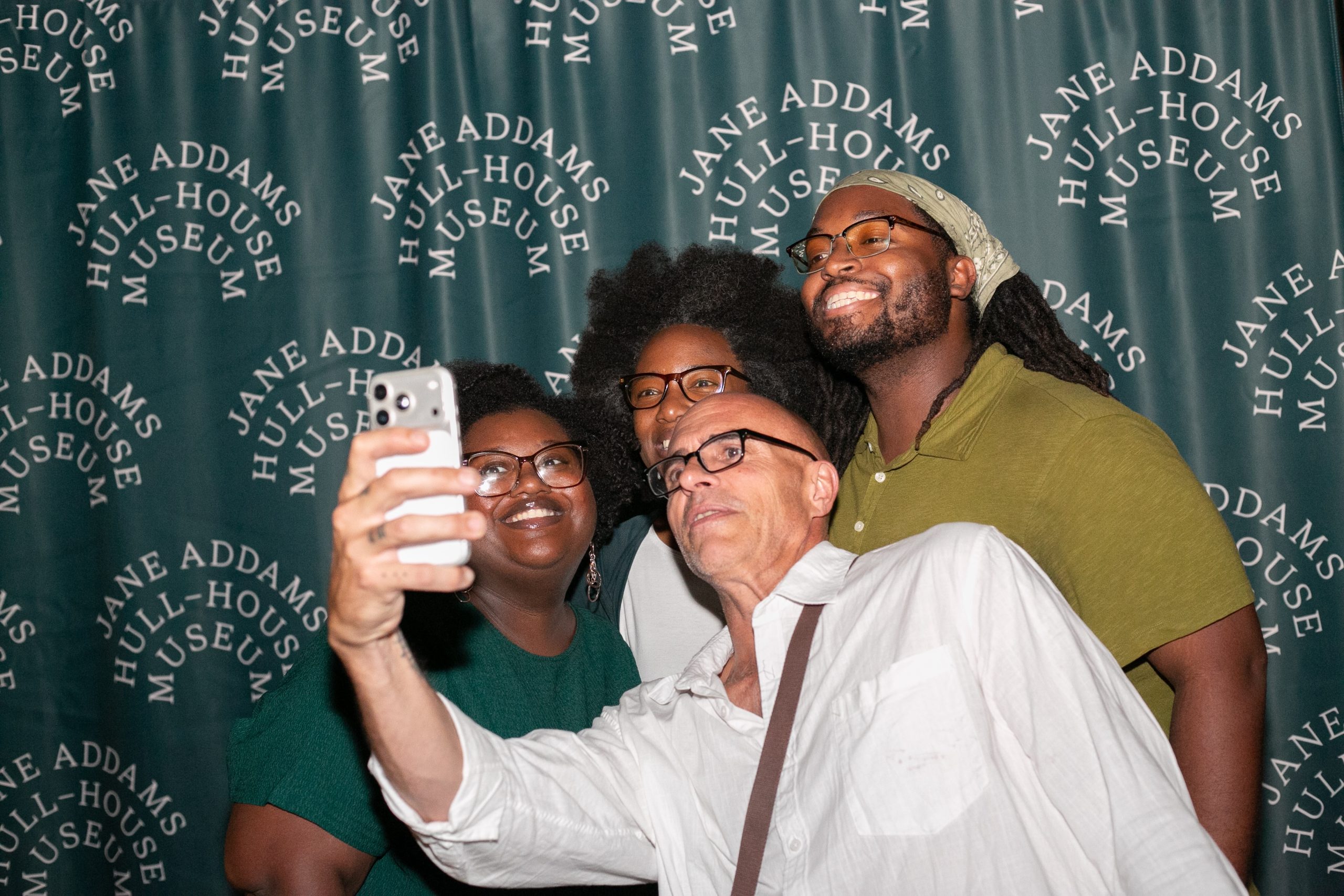Four people pose for a selfie in front of a green Jane Addams Hull-House Museum step and repeat banner.