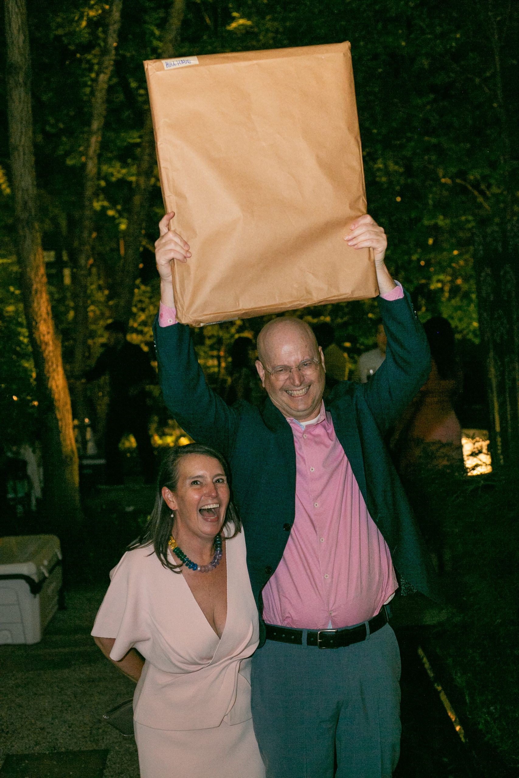A smiling man in a blazer and a woman in a dress hold a large, flat, brown paper-wrapped package high above their heads at an outdoor evening event.