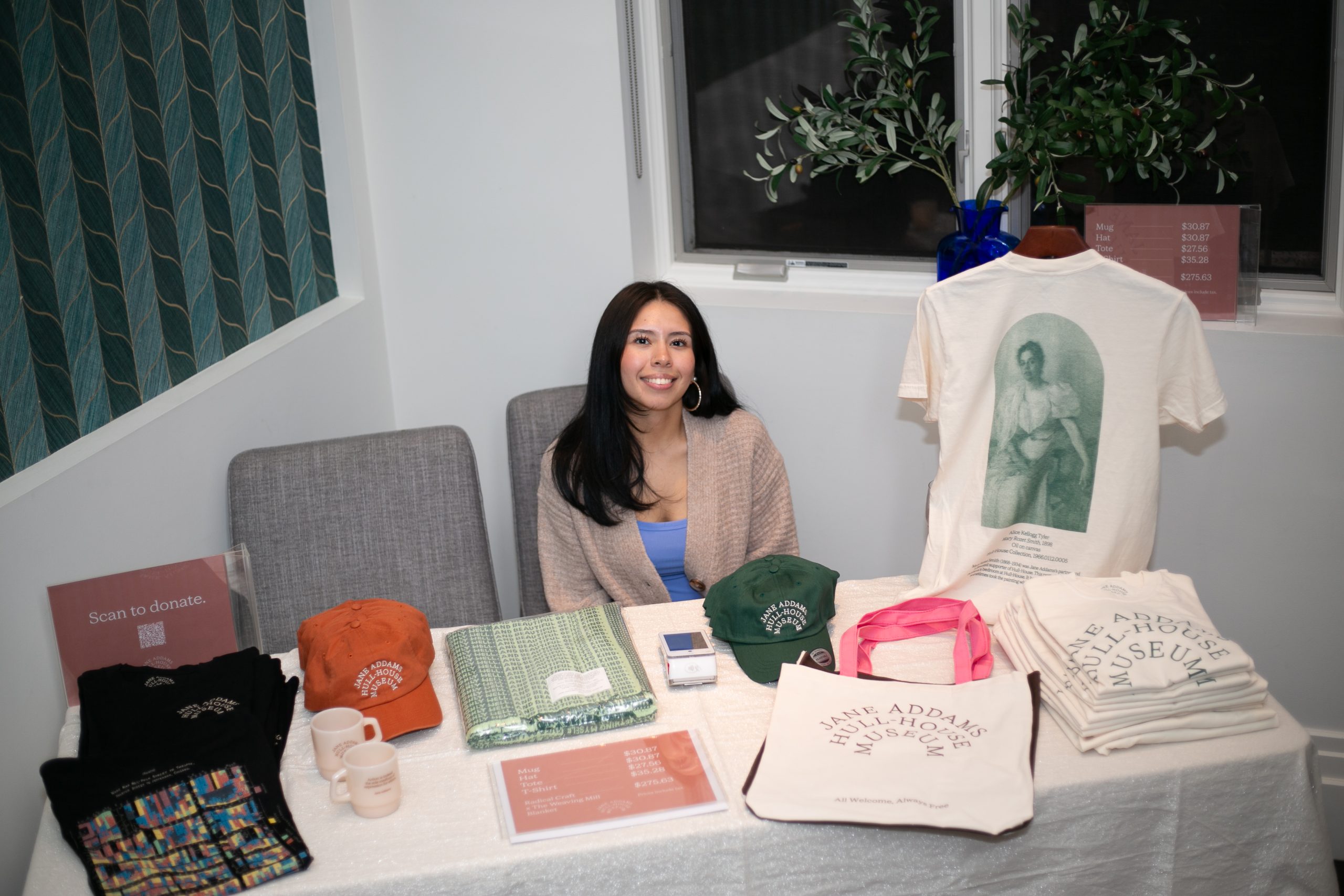 A woman smiles behind a merchandise table for the Jane Addams Hull-House Museum, which displays T-shirts, hats, tote bags, and mugs.