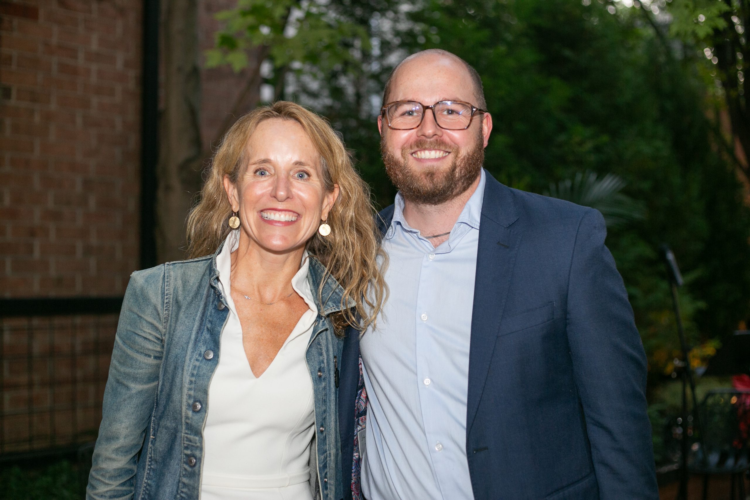 A smiling woman in a denim jacket and a smiling man in a navy blue blazer stand together outdoors.