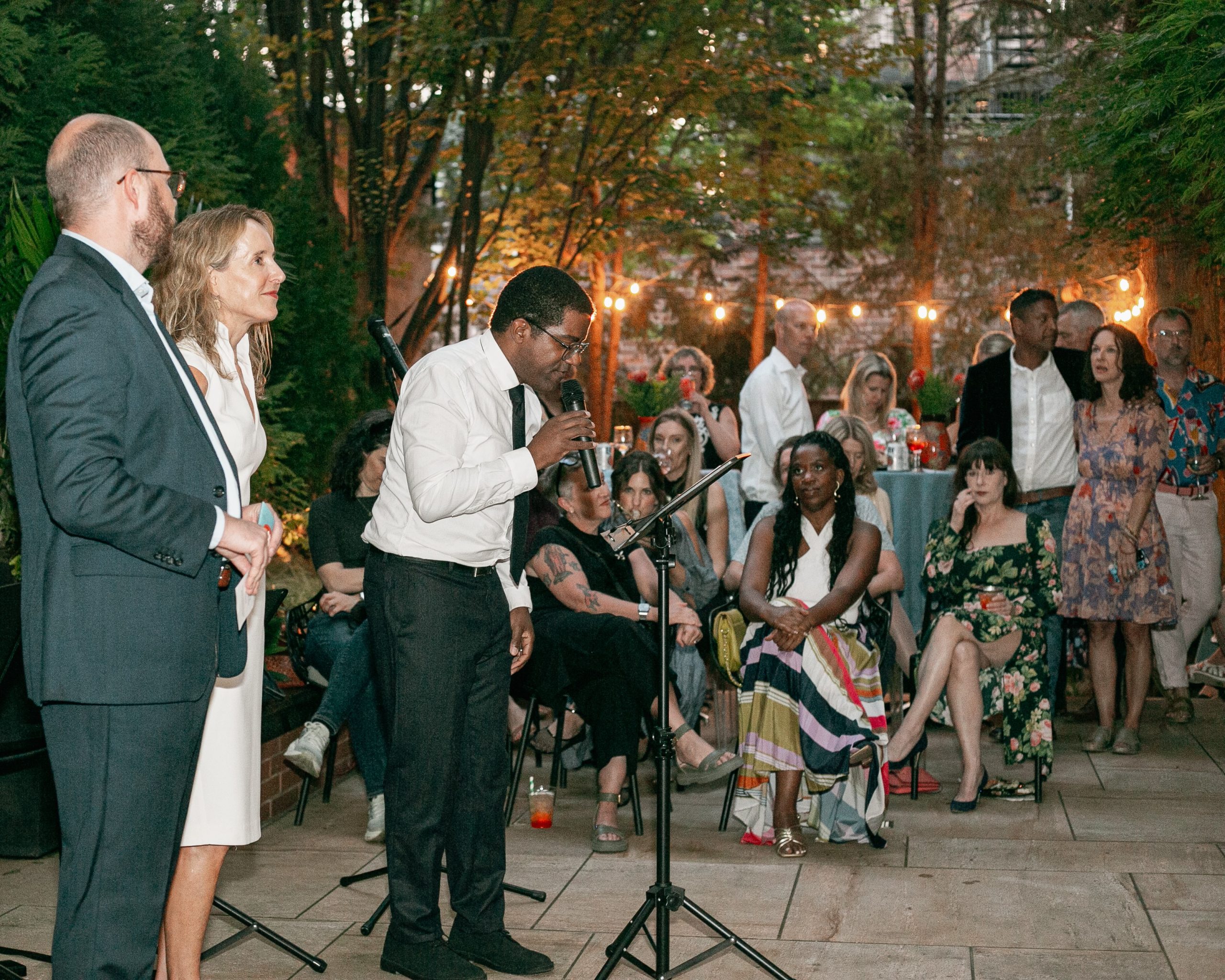 A man speaks into a microphone at an outdoor evening gathering, with a man and woman standing beside him and guests seated and standing in the background.