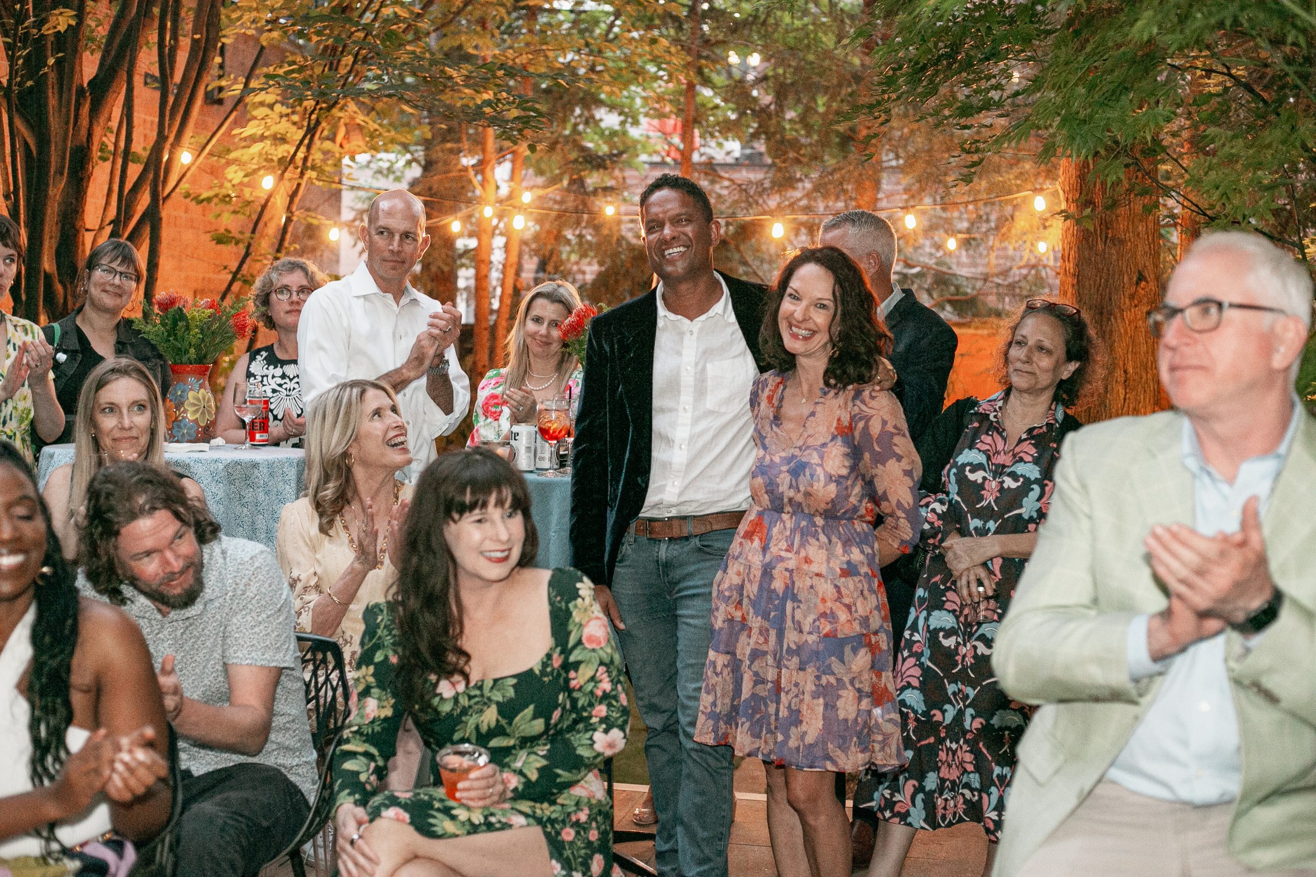 A group of people gathered at an outdoor evening party, with a man and woman standing in the center smiling while others around them applaud.