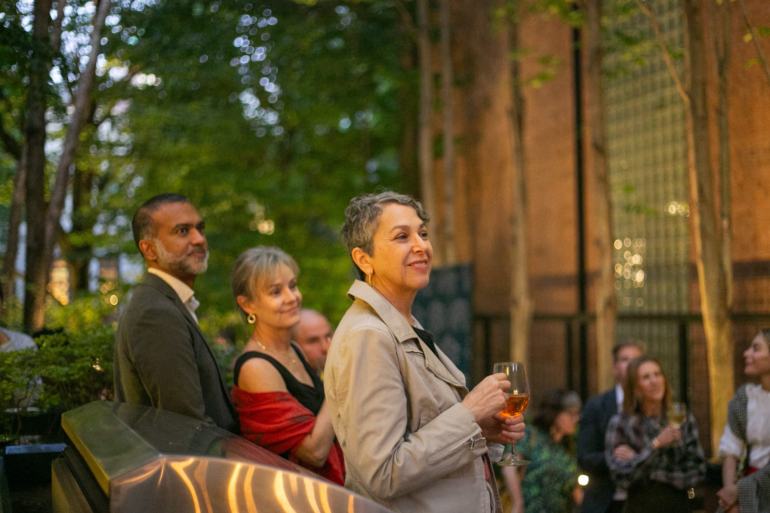 A group of people at an outdoor evening social event, with a woman in the foreground holding a glass of wine.