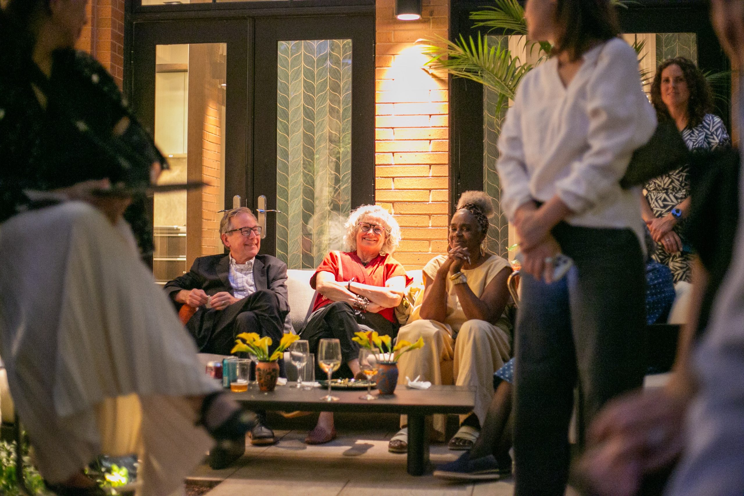 Three people sit on an outdoor patio sofa talking and smiling, while others stand nearby during an evening gathering.